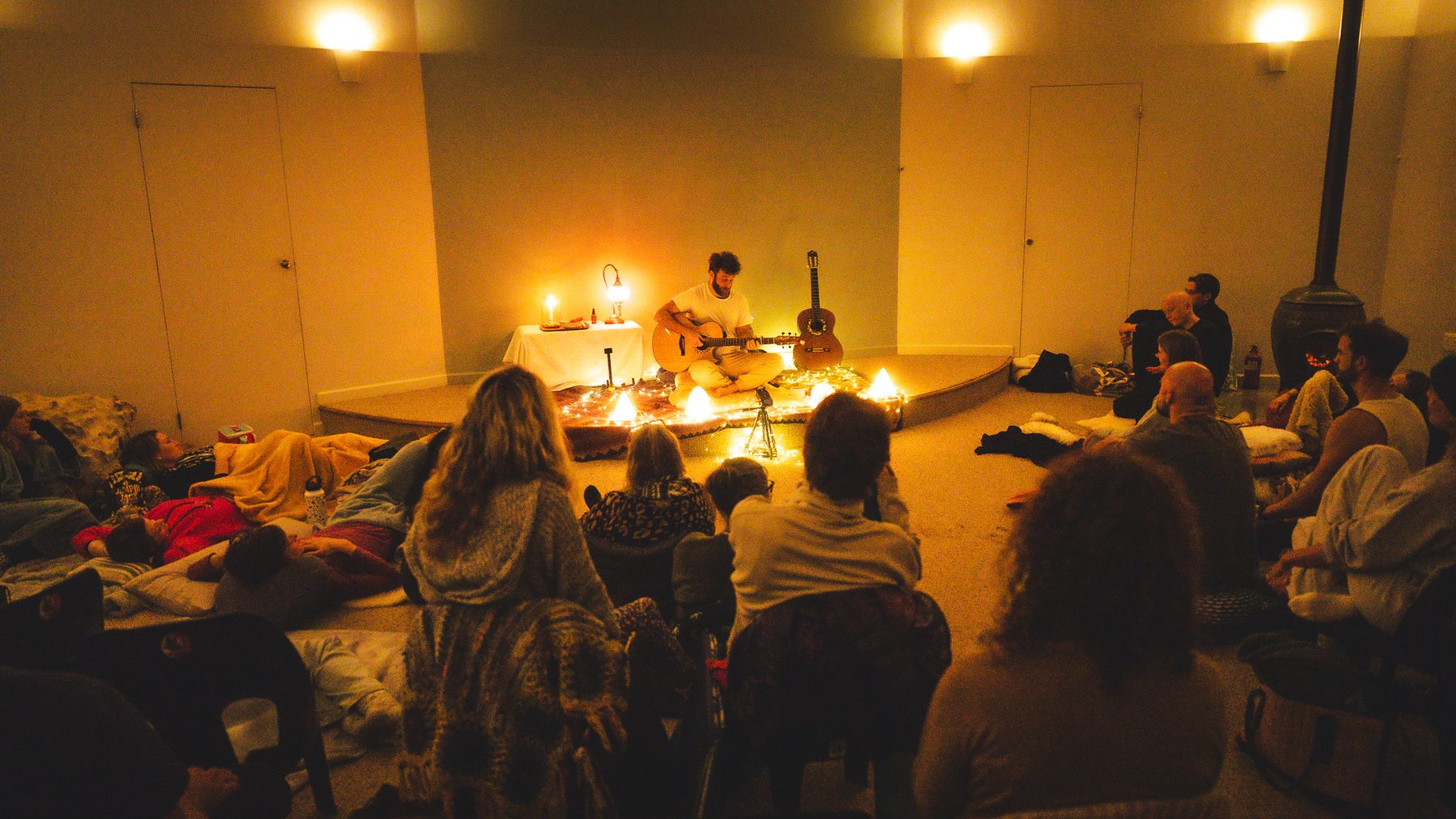 Nathan Maingard playing guitar by candlelight to a room of people lying and sitting around him