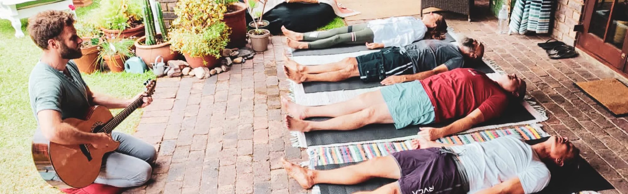 Nathan Maingard playing guitar beside a row of people resting in breathwork on a quiet patio